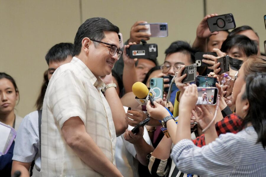 Senator Bam Aquino smiling while surrounded by a crowd of people and mobile phones during a press appearance.