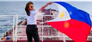 Second Officer KC Abigail Chin-Sood, a pioneering Mindanaoan seafarer, stands on the bridge deck of an international tanker vessel, confidently waving the Philippine flag to symbolize her successful 12-year career and the growing presence of Filipino women in maritime leadership.