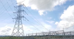 A high-voltage steel transmission tower and electrical pylon connecting lines to an expansive power substation under a blue sky with white clouds.
