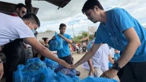 Aboitiz Foods personnel handing cardboard boxes of bottled water and plastic-wrapped food packs to residents standing in line at an outdoor disaster relief distribution site.