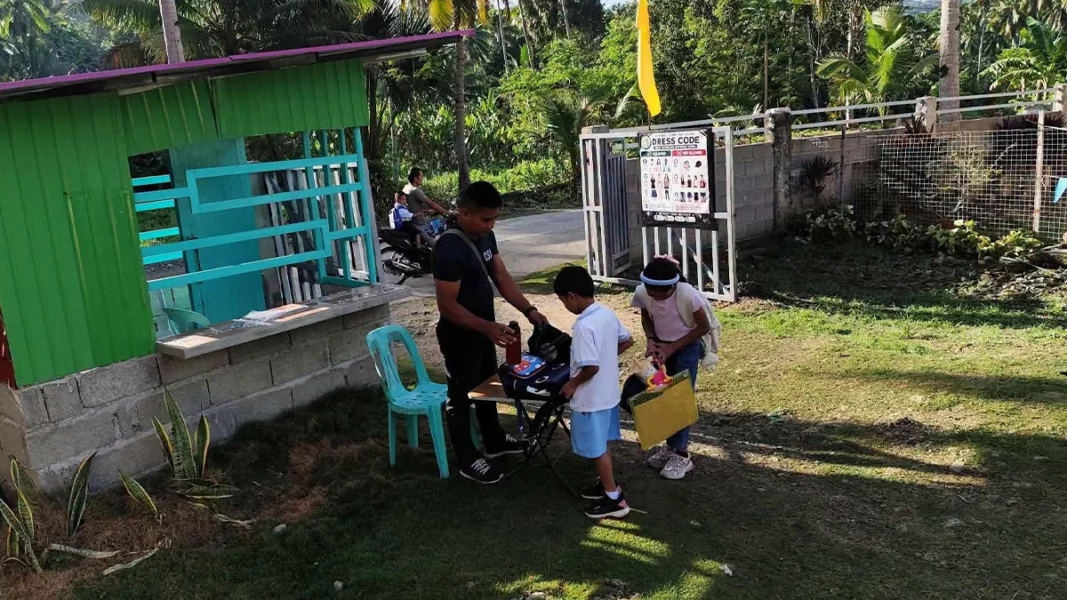 A campus security guard conducts a bag inspection and verifies identification cards at a public school entrance gate in Davao City.