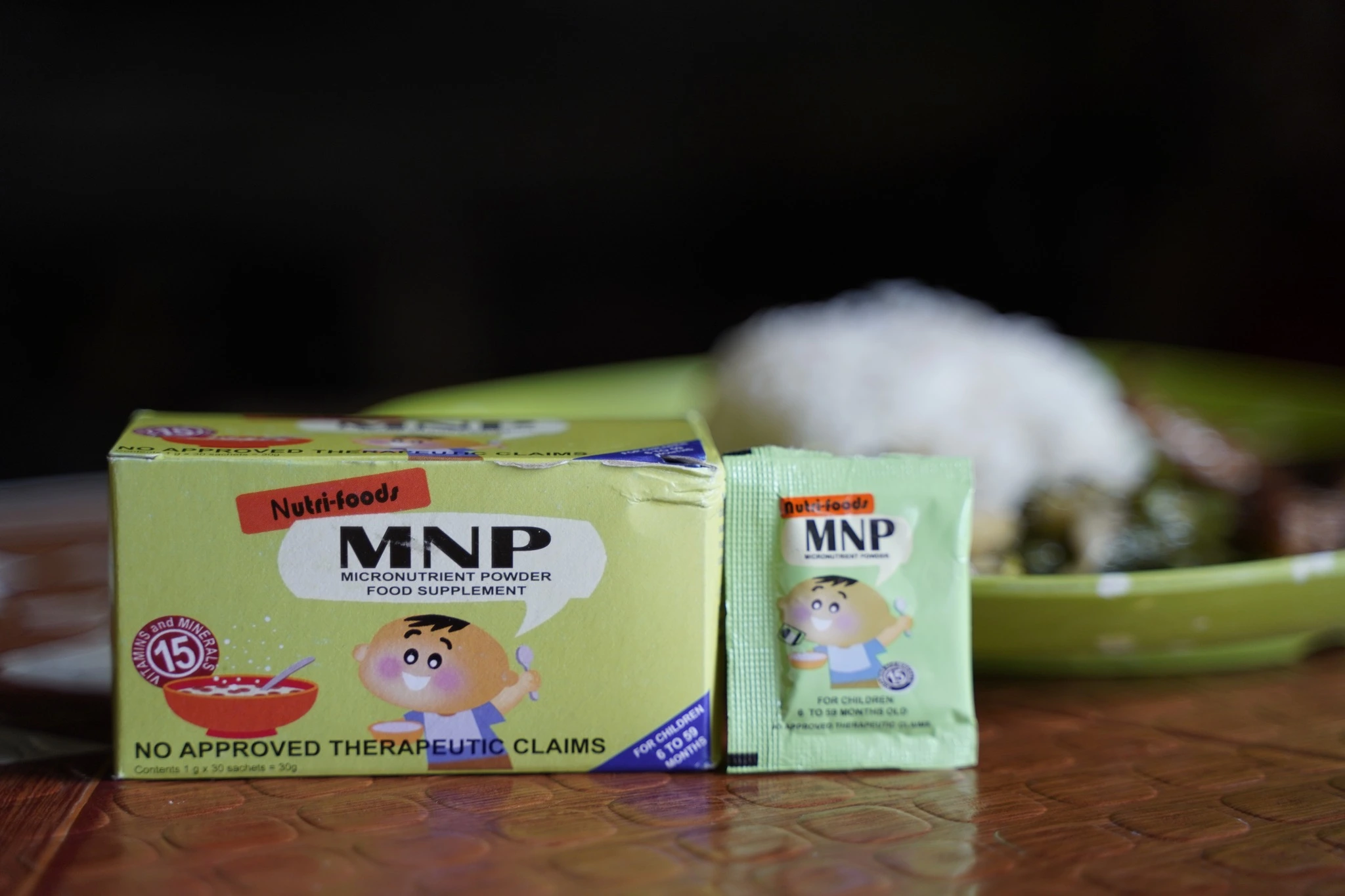 A box and an individual sachet of Nutri-foods Micronutrient Powder (MNP) food supplement for children aged 6 to 59 months, placed on a table with a meal blurred in the background.