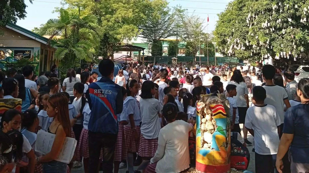 A group of school children and parents stand gathered together in an open outdoor courtyard during a safety evacuation in Iligan City following a major earthquake.