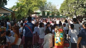 A group of school children and parents stand gathered together in an open outdoor courtyard during a safety evacuation in Iligan City following a major earthquake.
