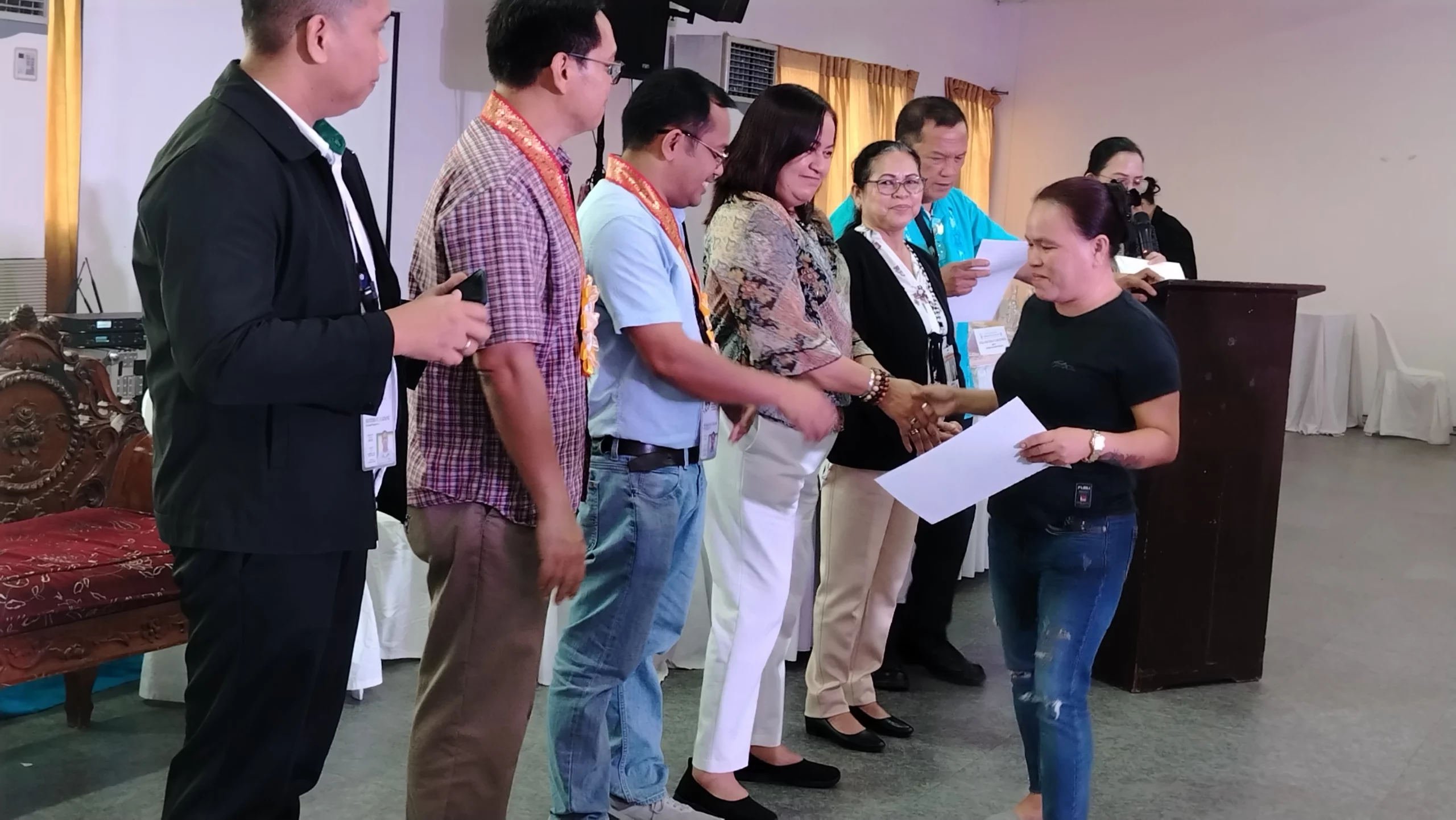 A row of local residents stands on a stage holding official printed certificates of award alongside government officials inside a crowded event hall.