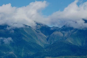 Lush green peaks of Mount Kalatungan, a large stratovolcano partially covered by thick white clouds under a blue sky in Bukidnon, Philippines.
