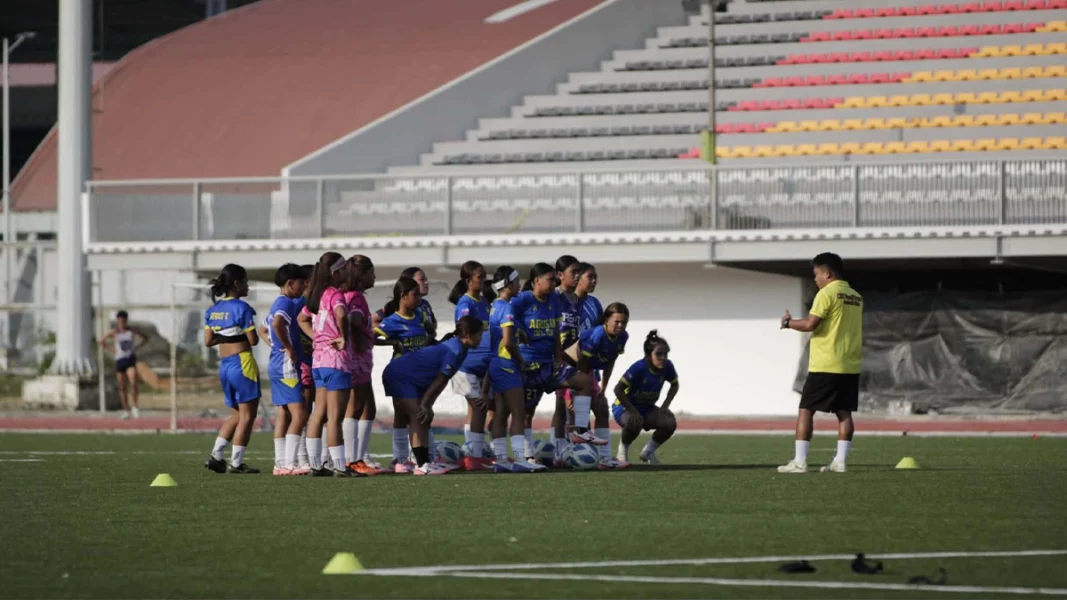 A youth female soccer team wearing blue and pink jerseys stands on a green turf field listening to their coach in a yellow shirt, with empty red and grey stadium grandstands in the background.