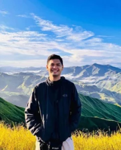 A portrait photo of a smiling, Alkharj Gomez Zamayla, wearing a black zip-up outdoor jacket with green mountain ridges and a cloudy blue sky in the background.