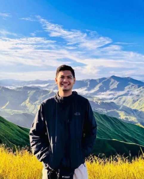 A portrait photo of a smiling, Alkharj Gomez Zamayla, wearing a black zip-up outdoor jacket with green mountain ridges and a cloudy blue sky in the background.