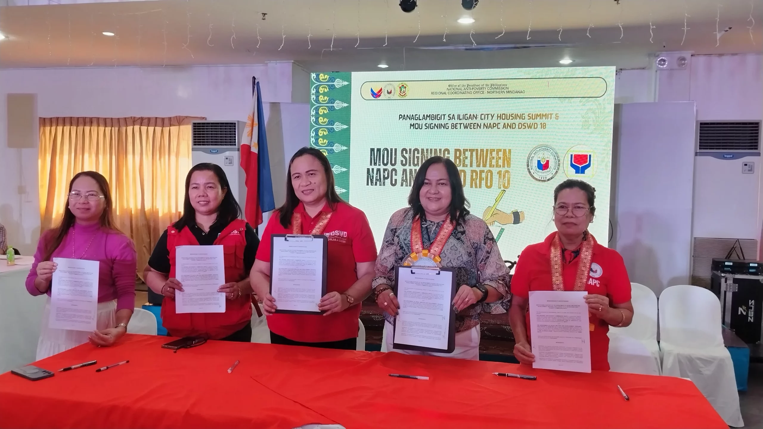 Five Officials stand side by side at a conference table showing the signed documents during an institutional summit in an indoor event hall.