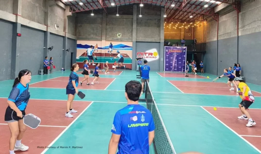 An indoor sports complex featuring multiple painted pickleball courts where men and women play doubles matches during a tournament at Nitro Pickleyard, with an official umpire in a blue jersey standing in the foreground.