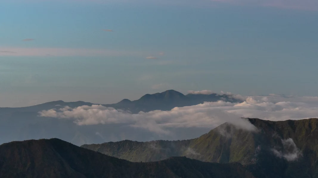 A wide, high-angle landscape view of the rugged, dark green ridges and peaks of the Mount Kalatungan mountain range in Bukidnon, with thick white layers of low-lying clouds and mist settling between the valleys under a pale blue and grey sky.