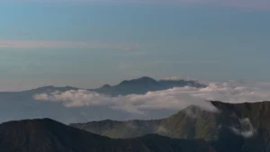 A wide, high-angle landscape view of the rugged, dark green ridges and peaks of the Mount Kalatungan mountain range in Bukidnon, with thick white layers of low-lying clouds and mist settling between the valleys under a pale blue and grey sky.