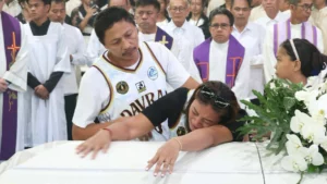 Rovelyn Baterbonia weeps while leaning over the white casket of her son, late student-athlete Bobet Baterbonia, as her husband, Rene Baterbonia Sr., places a hand on her shoulder to comfort her during a funeral service surrounded by Catholic clergy and mourners in Talacogon, Agusan del Sur.