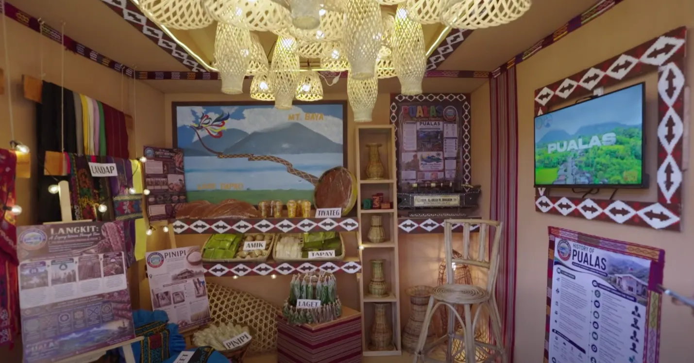 A cultural exhibition booth representing Pualas, Lanao del Sur, showcases traditional Maranao textiles, local delicacies, and regional handicrafts during a provincial heritage festival on July 8, 2026. The display features handwoven langkit fabrics, native agricultural products, and artwork depicting Mt. Baya and Lake Dapao to promote localized eco-tourism. (Screengrabbed from the Lanao del Sur Provincial Tourism Office)