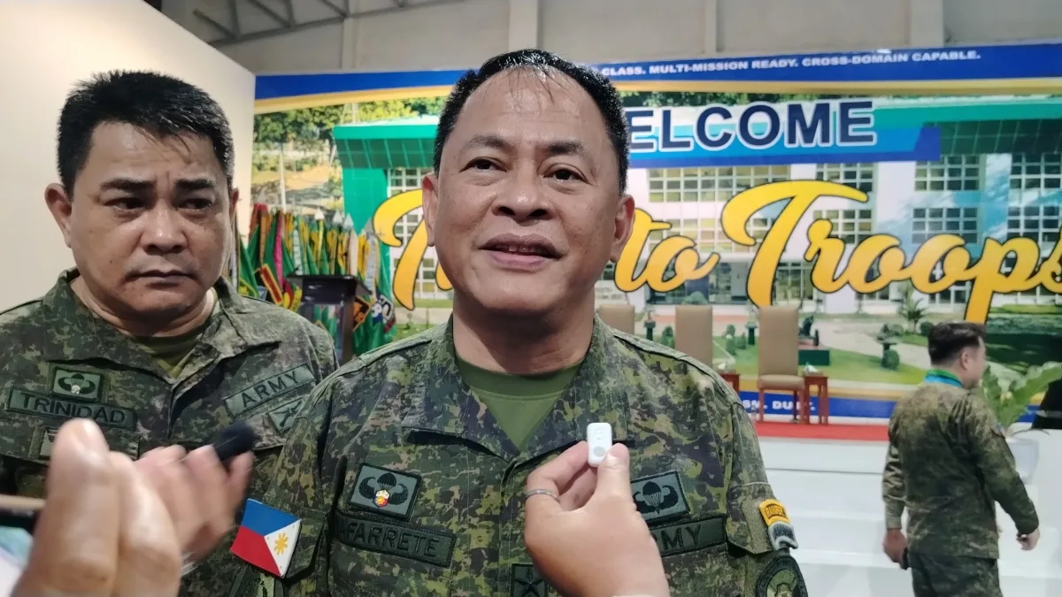 A uniformed Philippine Army officer speaks to members of the media while another officer stands beside him. Several military personnel and Philippine flags are visible inside a military facility.