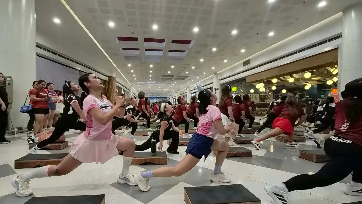 Participants in activewear execute step-dance lunges using raised wooden step platforms in a spacious, brightly lit mall atrium.