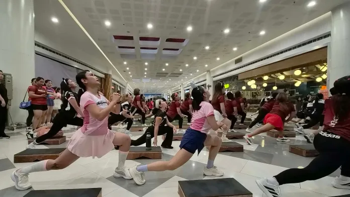 Participants in activewear execute step-dance lunges using raised wooden step platforms in a spacious, brightly lit mall atrium.