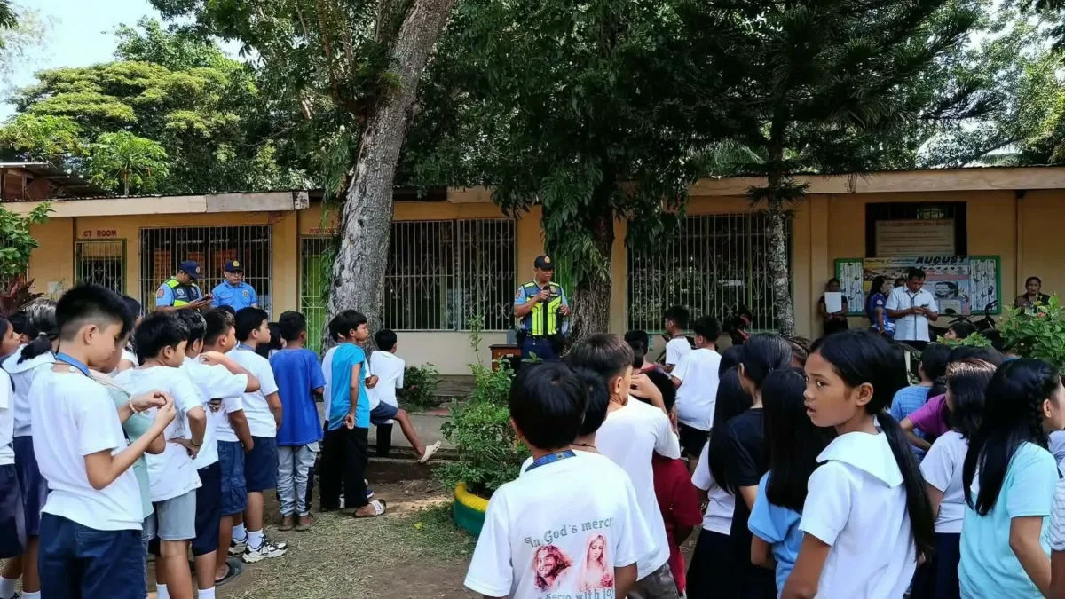 A police officer in a yellow high-visibility vest speaks into a microphone while standing in an open schoolyard facing a large group of elementary school students gathered outdoors near a classroom building.