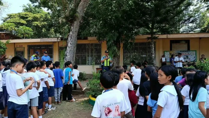 A police officer in a yellow high-visibility vest speaks into a microphone while standing in an open schoolyard facing a large group of elementary school students gathered outdoors near a classroom building.