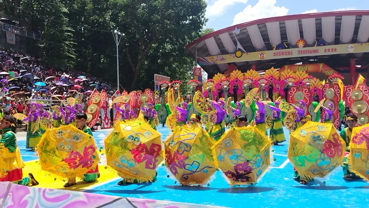 Performers in bright yellow and purple traditional costumes dance while holding decorated parasols on an outdoor blue stage during a festival competition. Spectators fill the grandstand under a sunny sky behind a banner reading Diyandi Festival 2026.