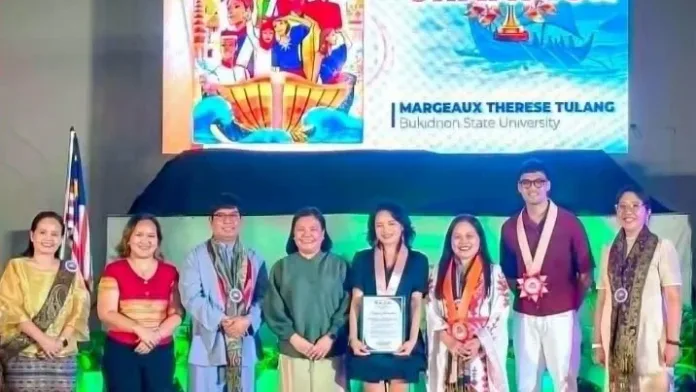 Margeaux Therese Tulang standing in the center holding her championship certificate, flanked by BukSU and CHED officials, with her winning poster and the word "CHAMPION" displayed on the screen behind her.