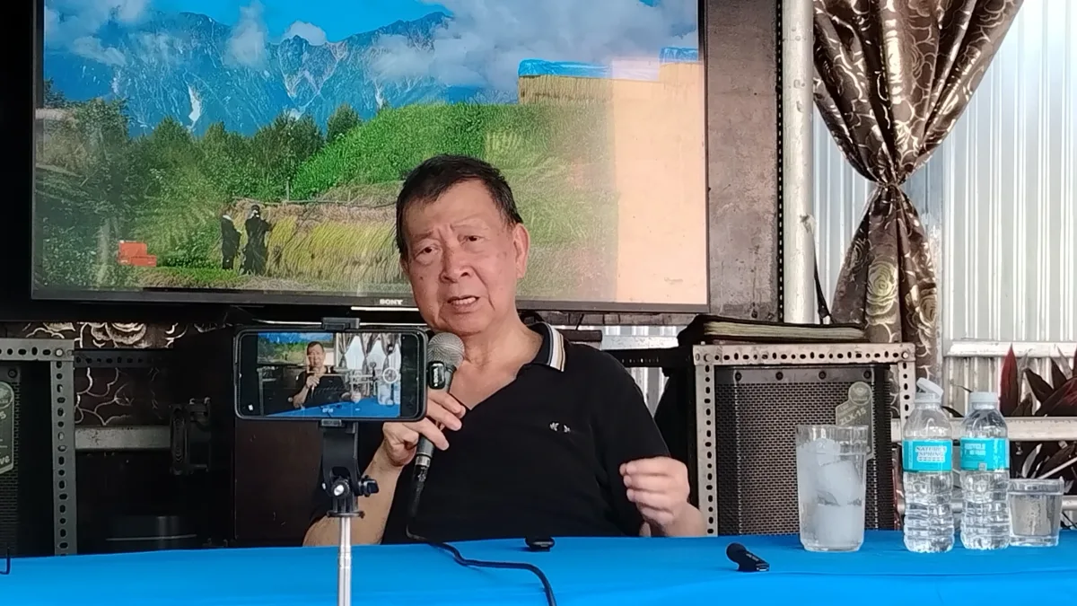 A man sitting at a blue-covered table speaks into a microphone held in his hand. A smartphone mounted on a mini tripod in front of him records the event, while a large television screen displaying a mountain landscape hangs on the wall behind him.