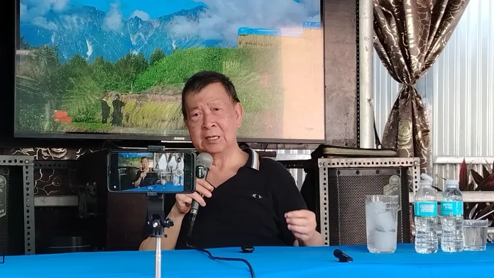 A man sitting at a blue-covered table speaks into a microphone held in his hand. A smartphone mounted on a mini tripod in front of him records the event, while a large television screen displaying a mountain landscape hangs on the wall behind him.