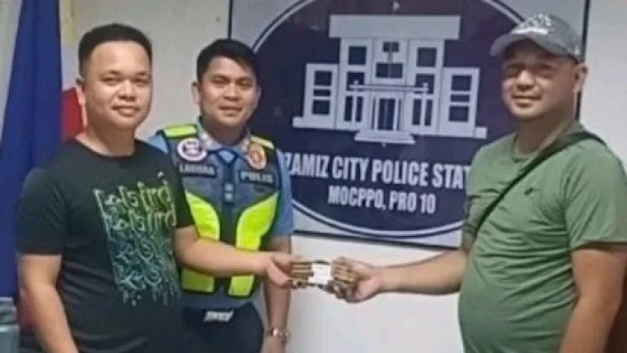 A job-order employee in a black shirt hands stacks of cash to a man wearing a green shirt and cap inside a police station, while a uniformed police officer stands in the background in front of the Ozamiz City Police Station seal.