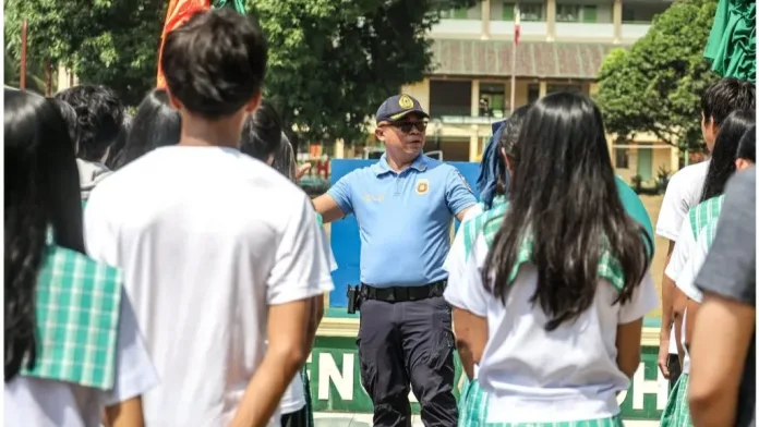A police officer in a blue uniform and cap speaks and gives directions to a group of high school students standing in formation outdoors. The students face the officer in front of a school building backdrop.