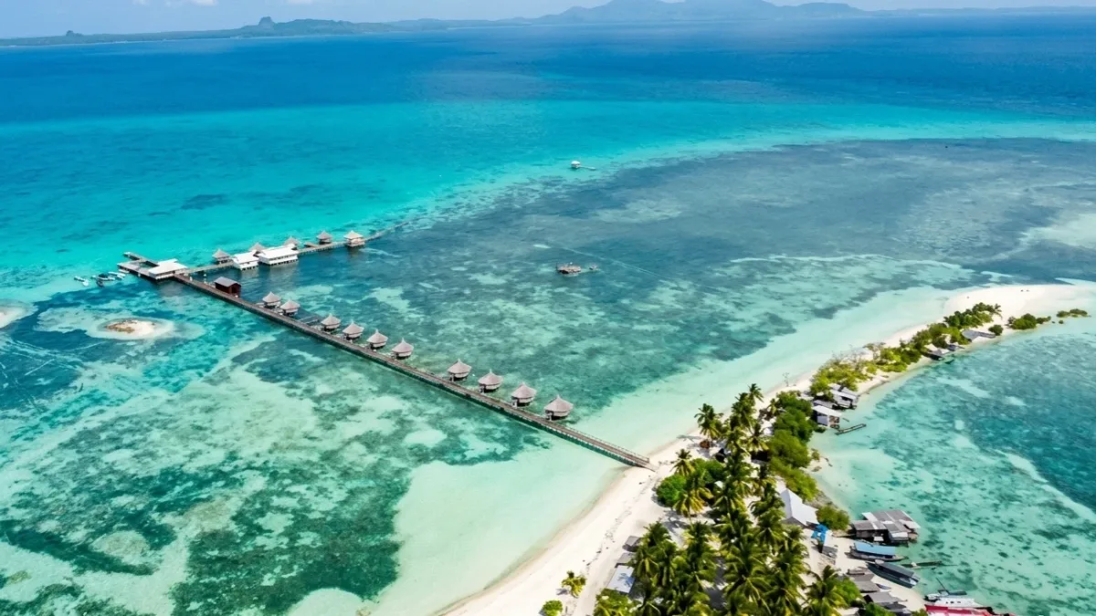 An aerial view of a tropical island resort surrounded by clear turquoise waters, white sand beaches, and lush coconut trees under a sunny sky. Wooden boardwalks and overwater stilt structures extend into the shallow sea.