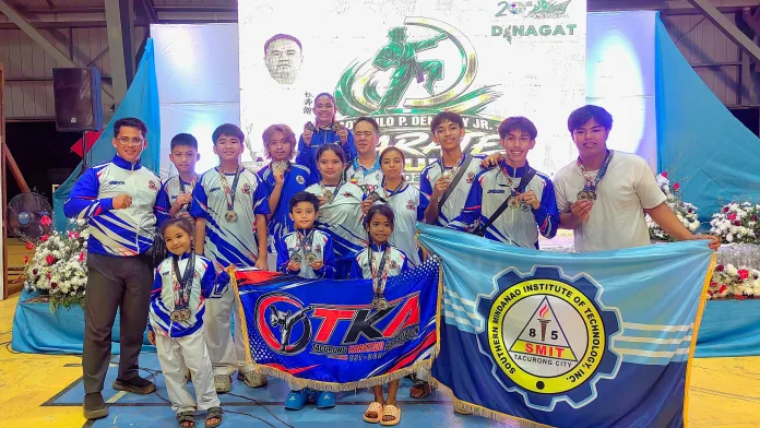 Members and coaches of the Tacurong Karatedo Association (TKA) pose with their medals, trophy banners, and school flag on stage during the 4th Governor Nilo P. Demerey Jr. Karate Cup 2026 in Dinagat Islands.