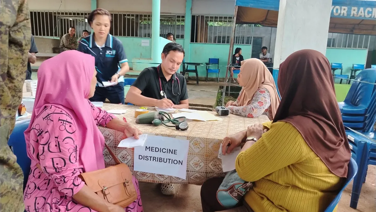 A medical volunteer writes notes at a table labeled "MEDICINE DISTRIBUTION" while attending to local women wearing traditional hijabs during a military community outreach program in Barangay Lininding, Munai.