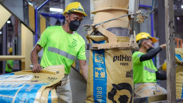 A factory worker wearing a bright green shirt, hard hat, and protective face mask guides a sack of Pilmico animal feed under an automated filling chute inside a feedmill facility.