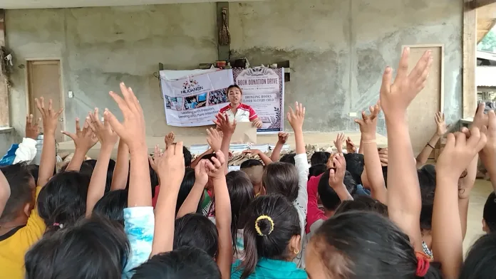 Higaonon youth leader Ahezel Llanes speaks from behind a stage table during a book donation drive and educational outreach program, as dozens of Indigenous learners eagerly raise their hands in a Barangay Rogongon community center in Iligan City.