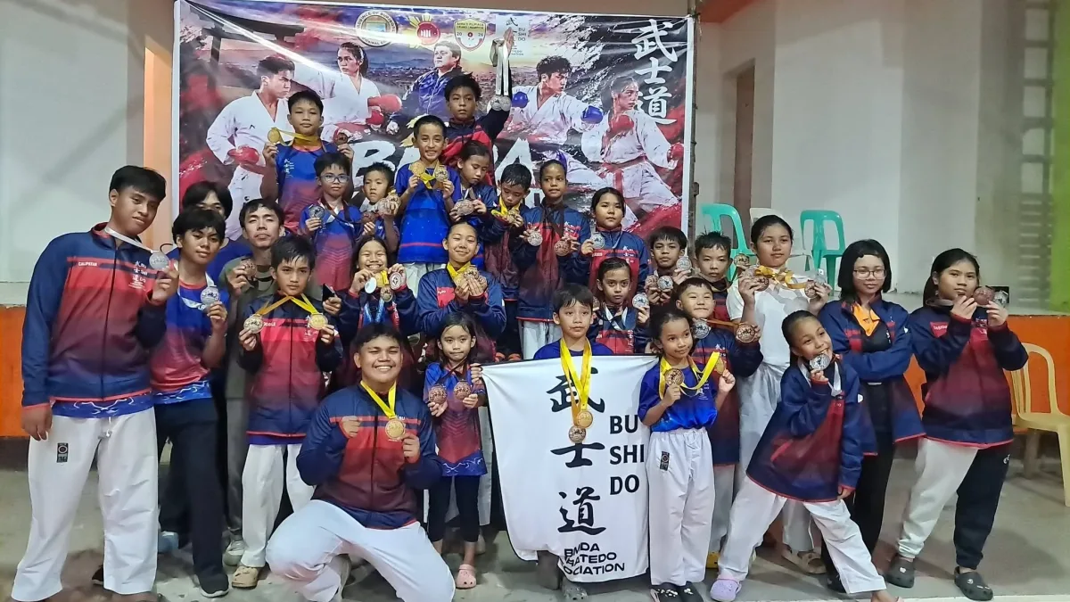 Young student-athletes, coaches, and officials of the Bushido Thru Martial Arts Development Association (BMDA) pose with their team banner while displaying competition medals in Malaybalay City.
