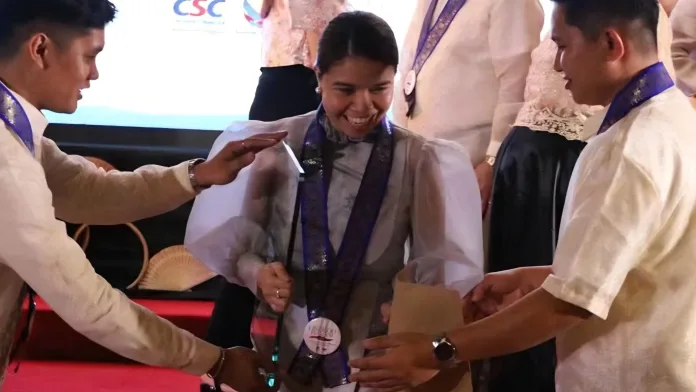 Xsyclyn Faith "Kanzai" B. Lumbatan smiling as she receives the CSC PRIME-HRM Bronze Award plaque on stage during the CSC Region 10 awarding ceremony.