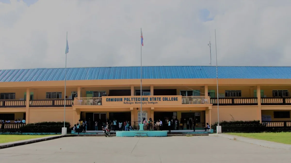 Exterior front facade of the main Camiguin Polytechnic State College building in Mambajao, Camiguin, featuring the Philippine flag and students gathered in the courtyard.