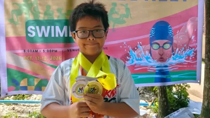 Eight-year-old swimmer Carl Fredrick Quizon wearing eyeglasses holds up three medals in front of the official DepEd 2026 Division Athletic Meet swimming backdrop at Kagay-an Lawndale Spring Resort in Cagayan de Oro City.