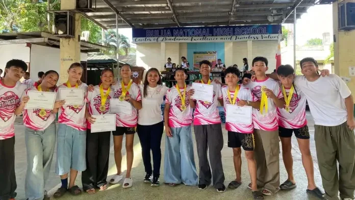 Student-athletes and coaches from Carmen National High School hold up certificates and medals under the covered court stage of Bulua National High School during the Cagayan de Oro Division Athletic Meet 2026.
