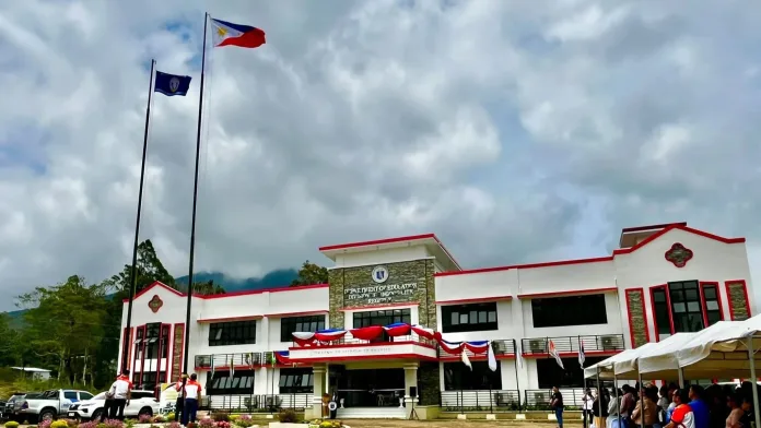 An exterior view of the newly constructed two-storey DepEd Gingoog City Division Office building with bunting decorations, flagpole, and attendees gathered under tents during the blessing ceremony.