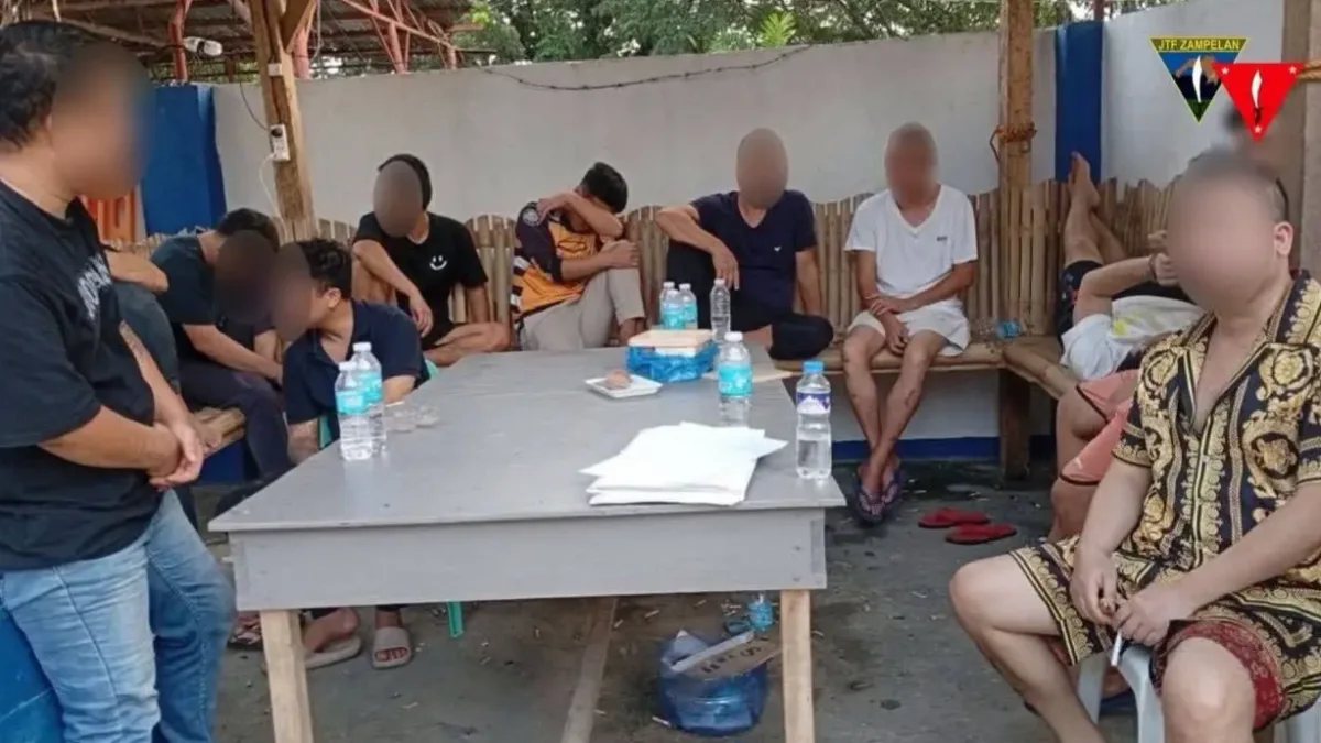 A group of Chinese nationals with blurred faces sit around wooden tables inside a facility in Zamboanga Sibugay while immigration and military intelligence officers process their documentation.