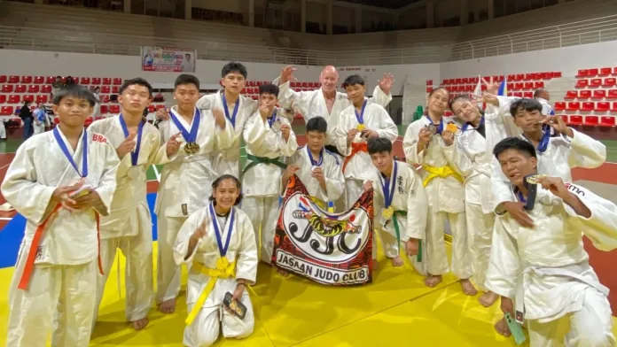 Members of the Jasaan Judo Club wearing white judogi pose enthusiastically on the mat while holding their medals, certificates, and team banner inside the gym in Zamboanga City.