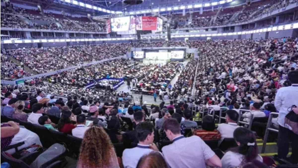 A wide, elevated view of thousands of attendees packing a multi-tiered indoor stadium arena during an international convention hosted by Jehovah's Witnesses.