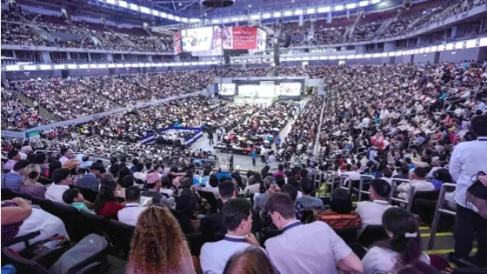 A wide, elevated view of thousands of attendees packing a multi-tiered indoor stadium arena during an international convention hosted by Jehovah's Witnesses.