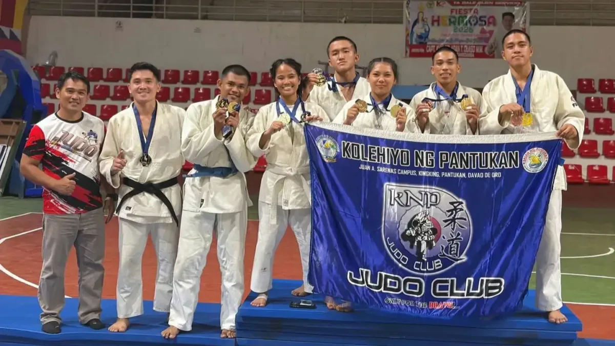 Seven judokas in white gis and their coach from Kolehiyo ng Pantukan pose on a podium mat holding their school banner and 13 tournament medals inside the Mayor Vitaliano D. Agan Coliseum in Zamboanga City.