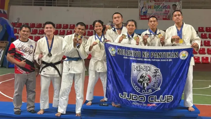 Seven judokas in white gis and their coach from Kolehiyo ng Pantukan pose on a podium mat holding their school banner and 13 tournament medals inside the Mayor Vitaliano D. Agan Coliseum in Zamboanga City.