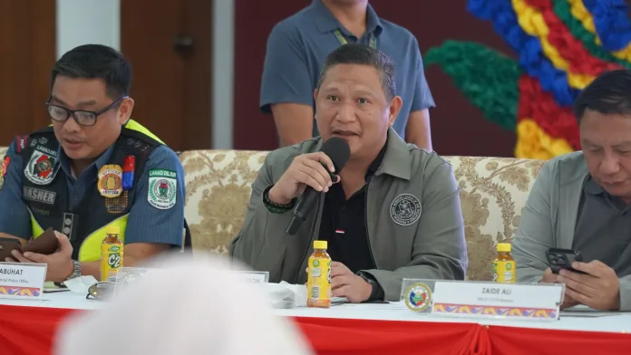 Lanao del Sur Provincial Board Member Saifullah Aldine Macarambon holding a microphone while presiding over the third-quarter joint meeting of the Provincial Peace and Order Council and Provincial Anti-Drug Abuse Council alongside police and local officials.