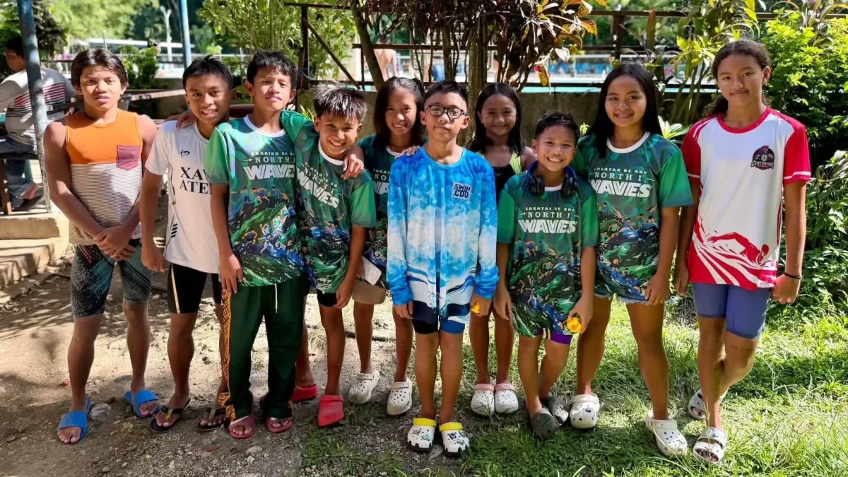 Young student-athletes from the Let's Swim CDO club pose together outdoors in athletic shirts, including "North I Waves" gear, following their qualification for the regional meet in Camiguin.