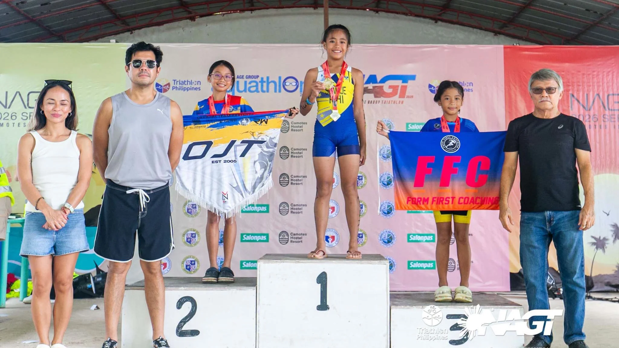 Nine-year-old triathlete Ma. Georjina Concepcion Pacquing Sumaje of Zamboanga City smiles on the top step of the winner's podium holding her gold medal, flanked by second-place finisher Maiko Aleno and third-place finisher Stacey Ailia Aisha Escala during the award ceremony at the 2026 National Age Group Triathlon Camotes Island Series in Cebu.
